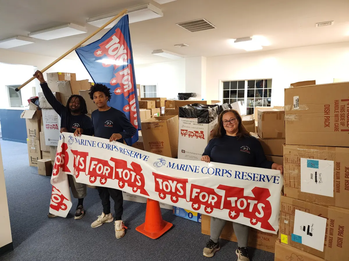 M.C. Movers crew holding a Toys for Tots banner and flag with donated toys collected for the Marine Corps Reserve drive